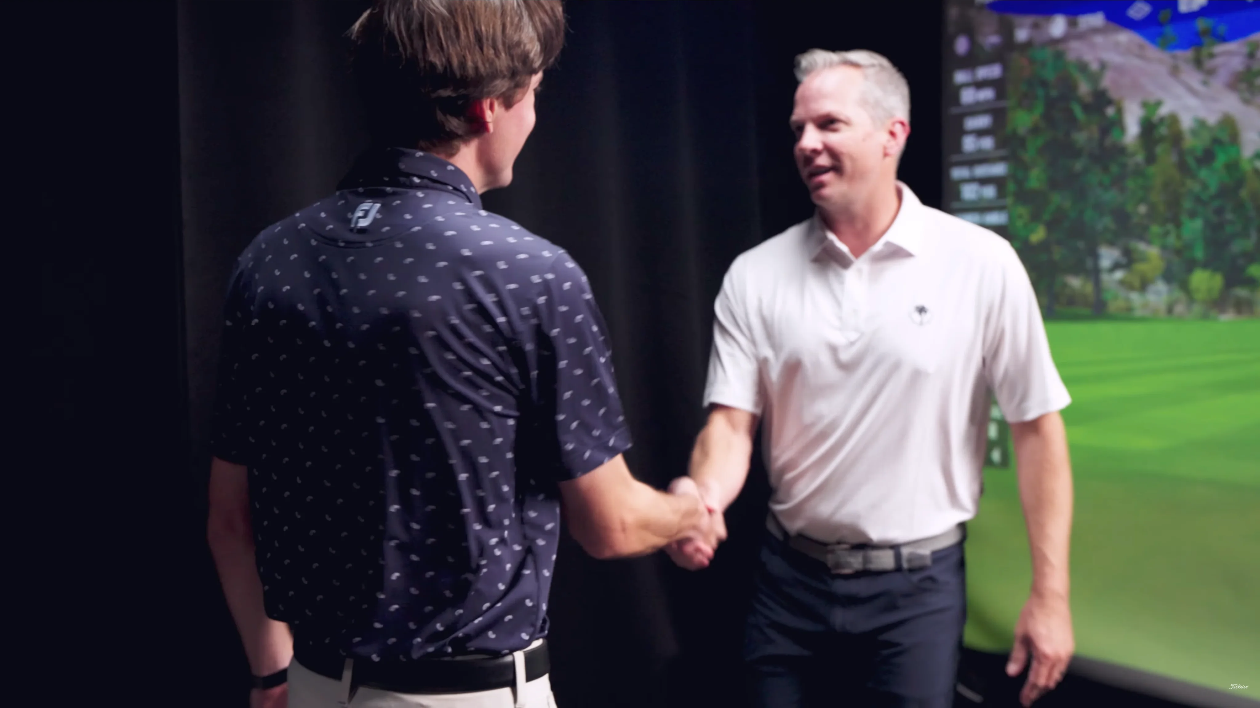 Two men shake hands in a golf simulator bay.