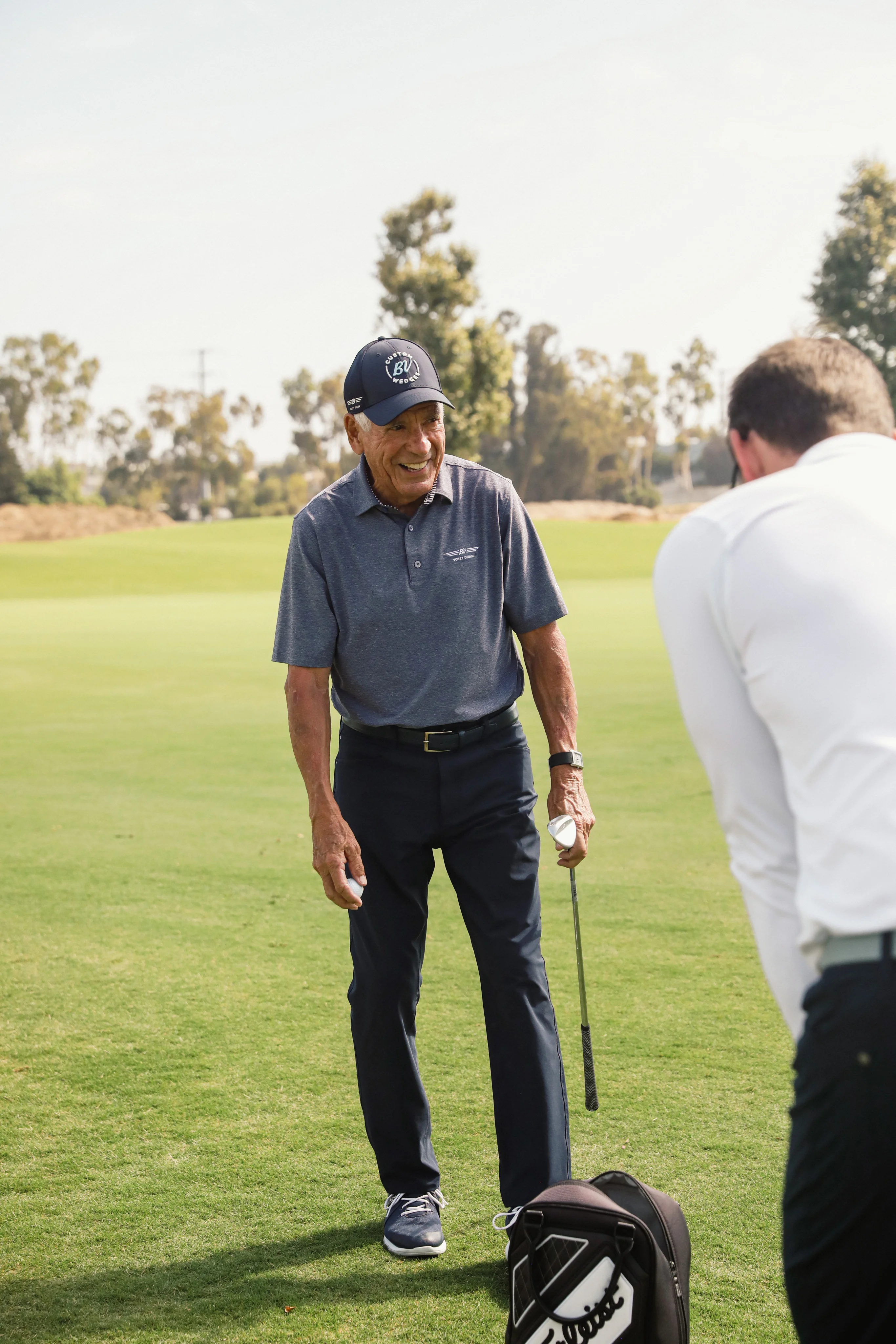 Bob Vokey smiles as he holds an golf club and golf ball on the course.