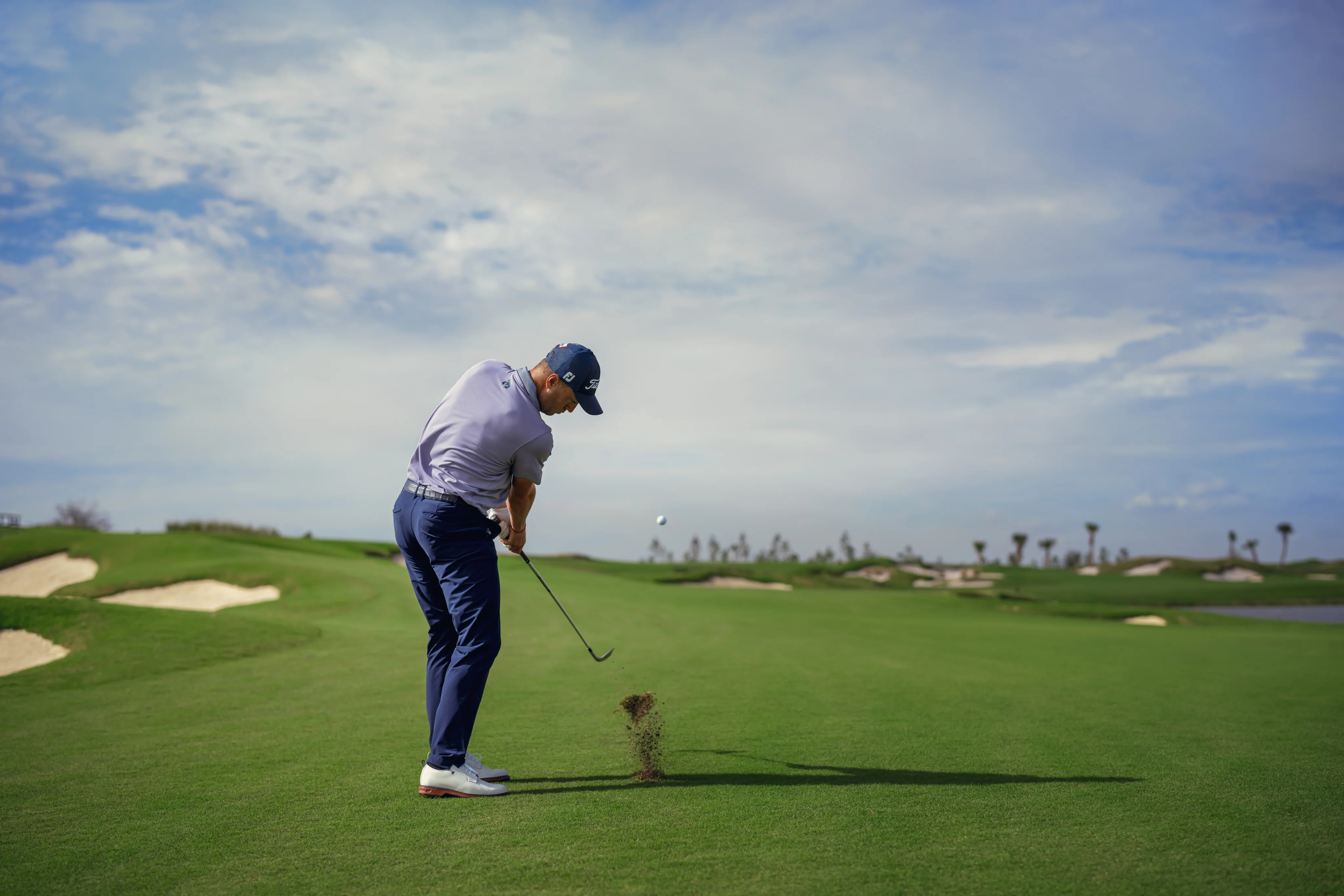 View from behind as a Justin Thomas strikes the golf ball with a golf club on the course.